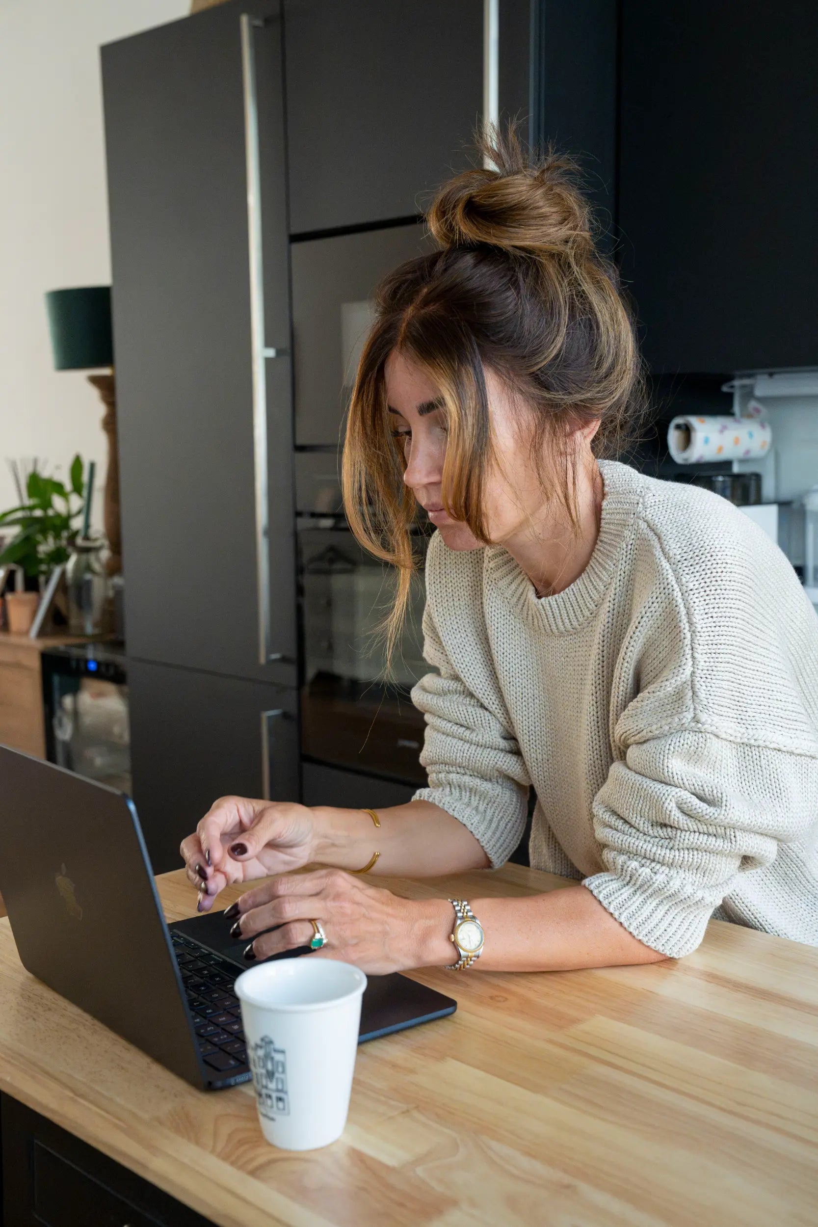 Woman with her hair in a top knot working on a laptop at a kitchen counter with a coffee cup