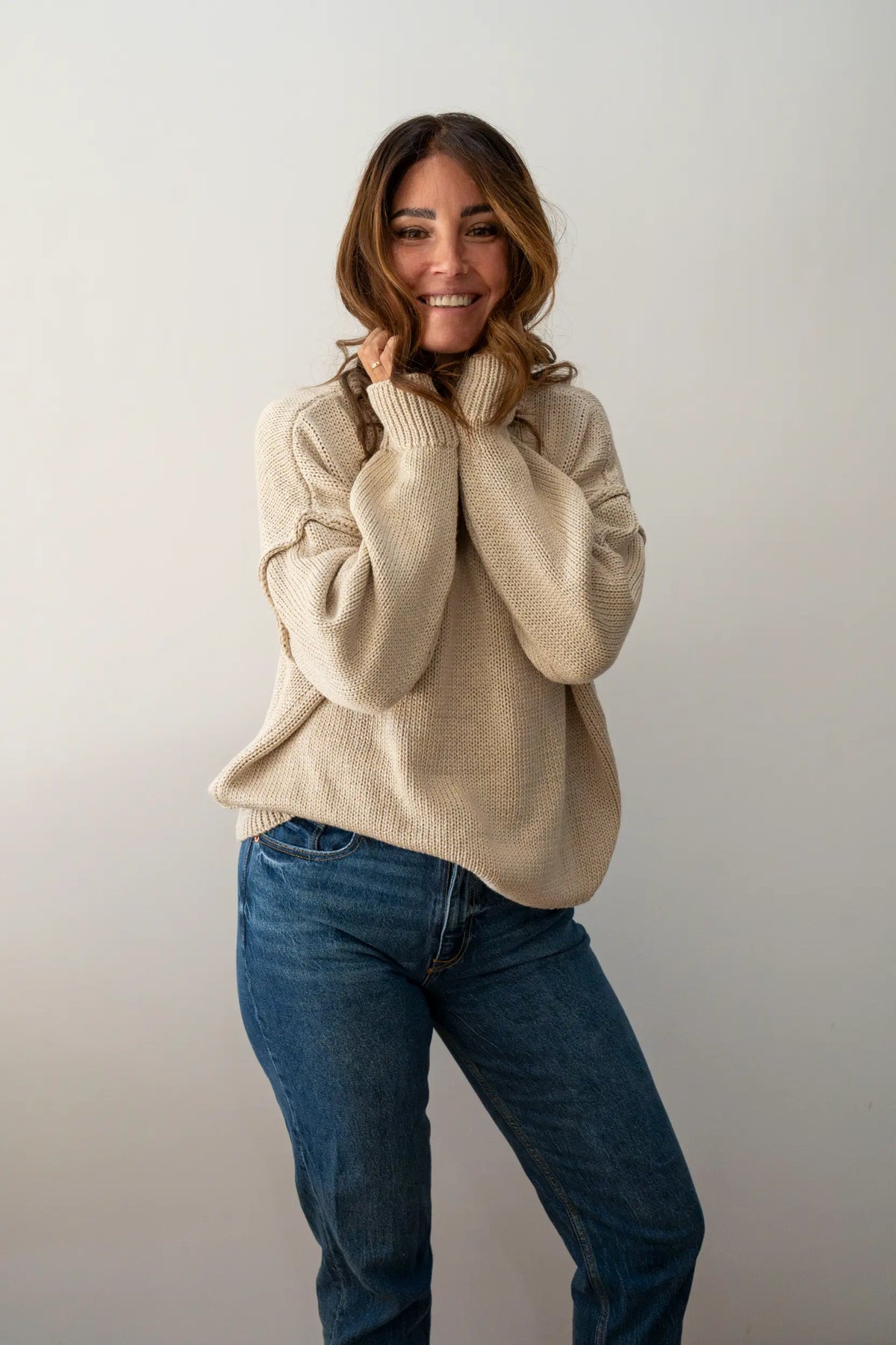 Smiling woman in a cream knit jumper and jeans wearing a wavy Bronzed Caramel human hair wig, full-length shot