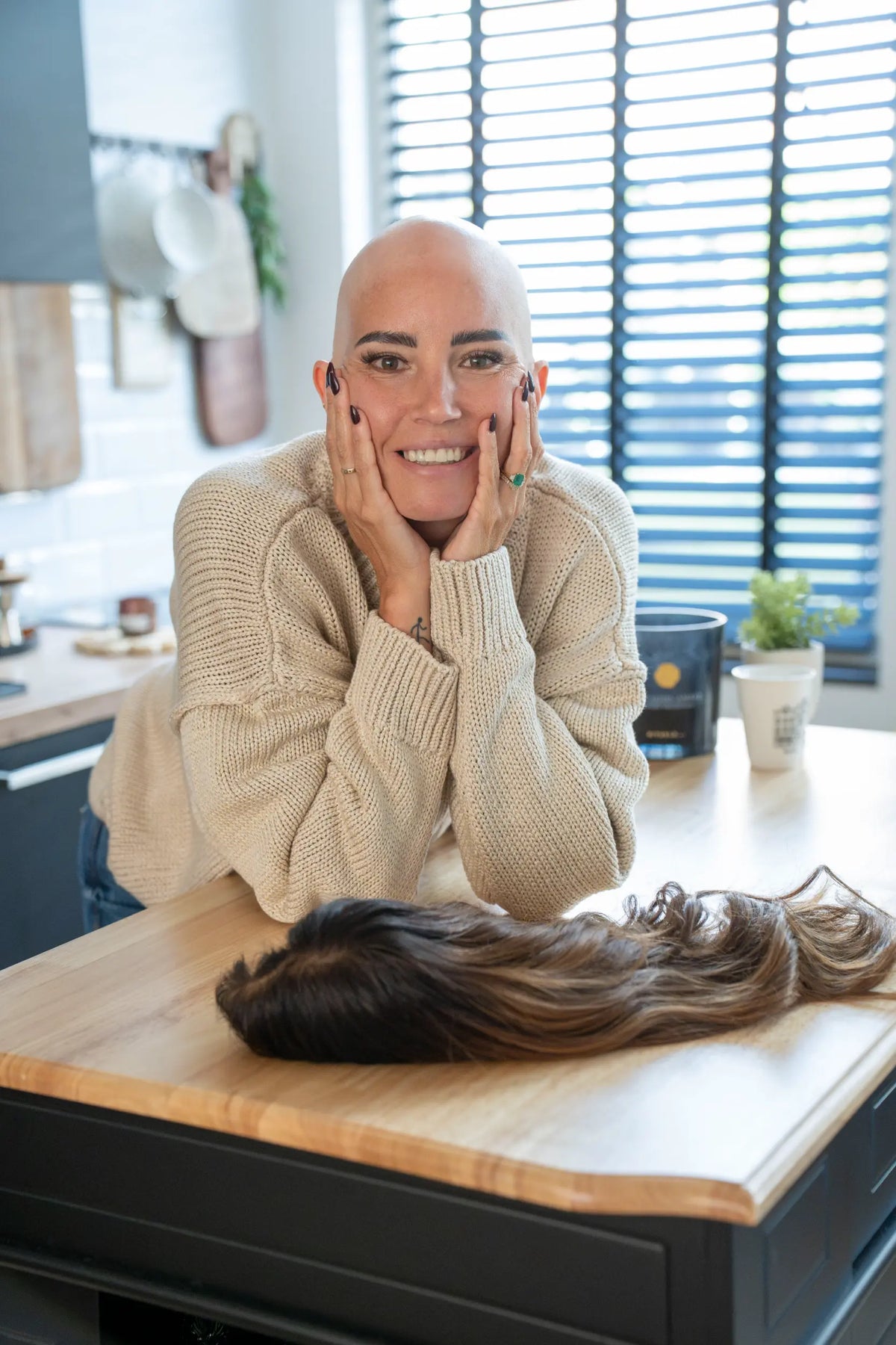 Natalie, founder of She Is Wearing It, who has alopecia, smiling at a kitchen counter beside a long wavy human hair wig